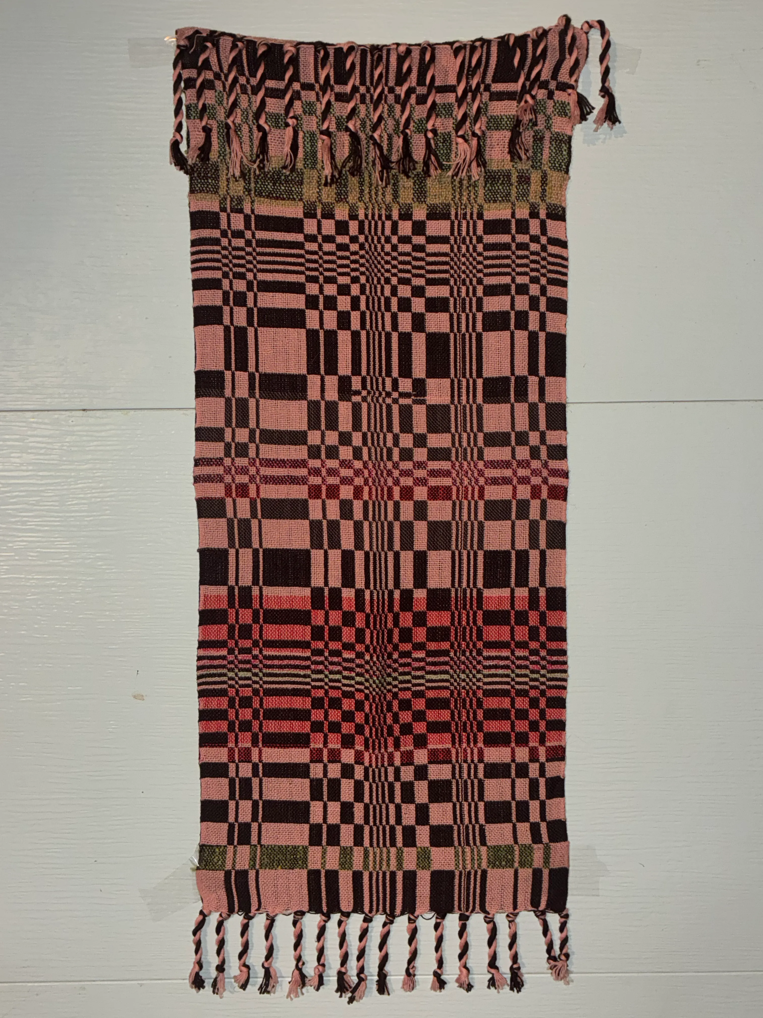 Rectangular woven textile laid flat on a white surface showing a complex double-cloth checkerboard pattern in pink, dark brown, and red, with twisted fringe along both short ends