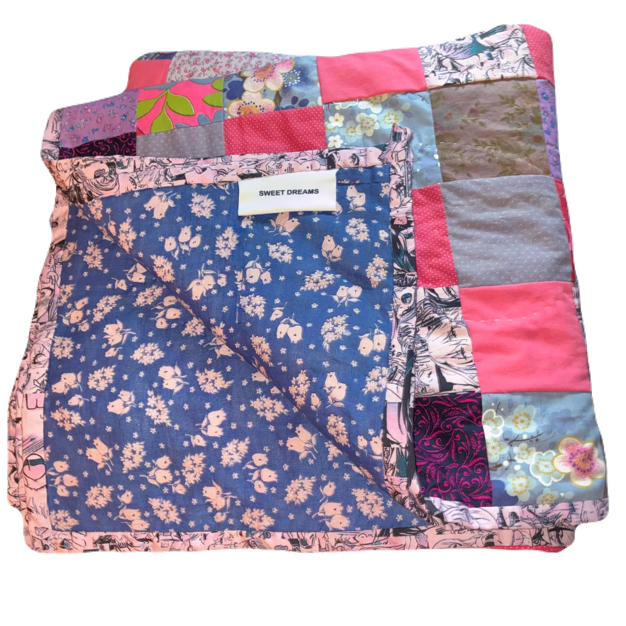 A folded patchwork quilt laid against a white background, showing a top made from varied printed cotton fabrics — blue floral, hot pink, lavender, teal, and multicolor patterns — with a SWEET DREAMS woven label, a pink floral border, and blue floral backing fabric folded over the edge
