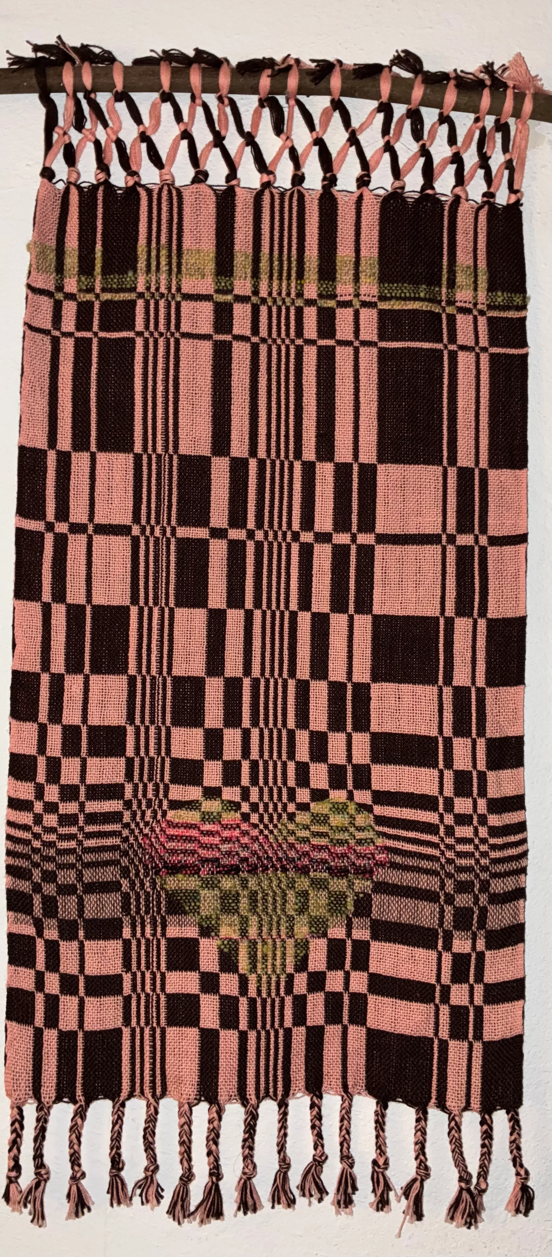 Woven textile hanging from a wooden branch against a white wall, showing a double-cloth pattern in pink and dark brown with a tapestry-woven heart motif in the lower center, with twisted fringe at the bottom