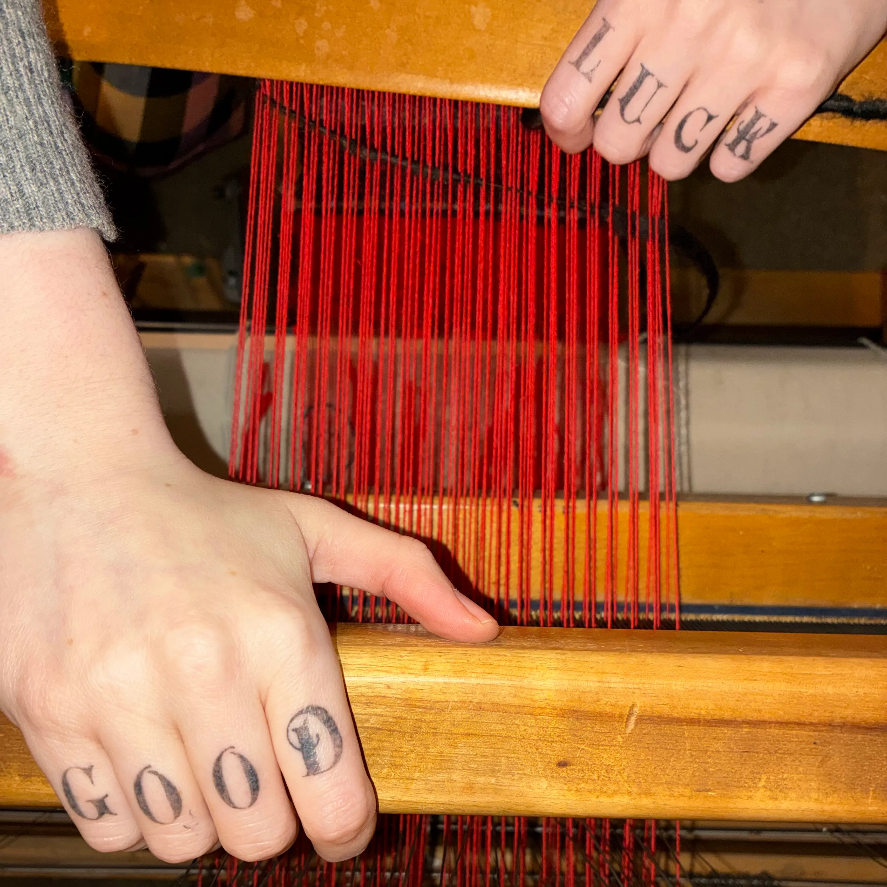 two hands threading a deep red warp on a wooden loom