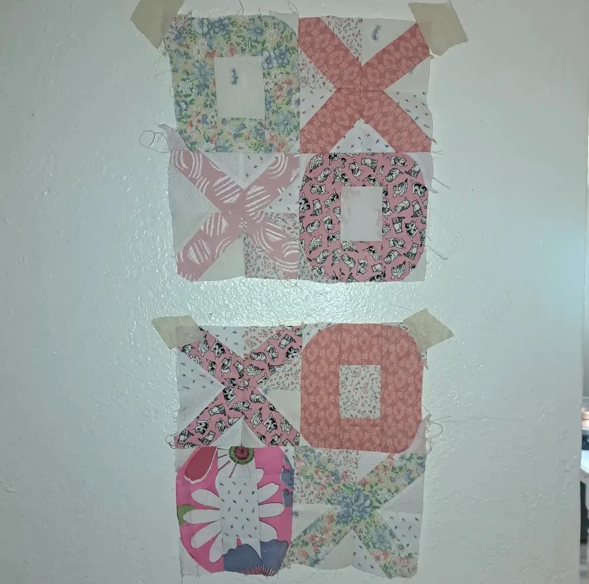 Four quilt blocks pinned to a white wall showing a hugs-and-kisses (XOXO) pattern in progress, with X and O forms pieced from pink, white, and floral repurposed cotton fabrics, raw edges visible, taped at the corners