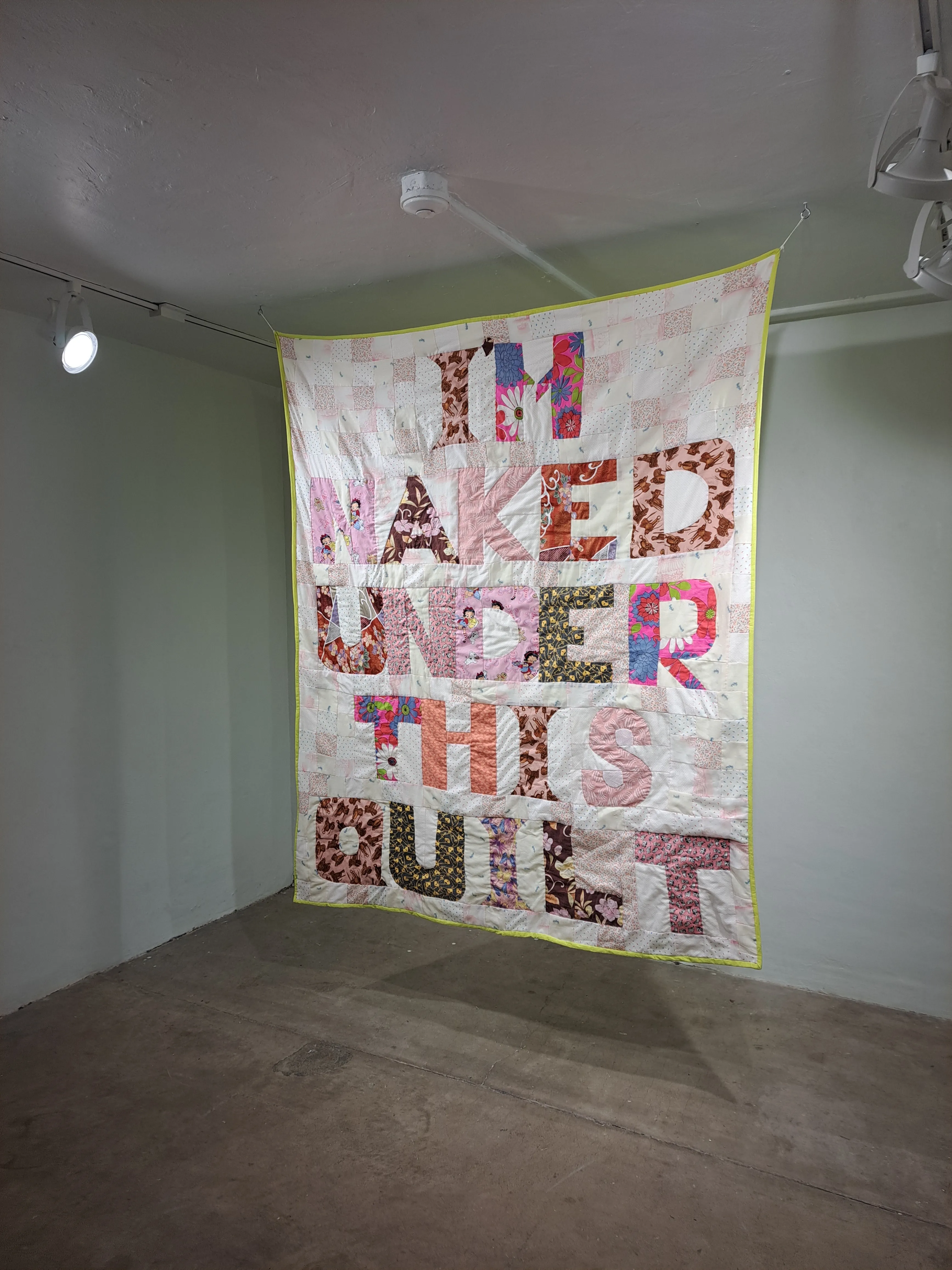A patchwork quilt hung from the ceiling of a gallery space, photographed from below at an angle, showing colorful letter forms cut from patterned fabrics — florals, animal prints, and geometric — in pink, orange, brown, and multicolor tones on a pale background with a yellow-green border