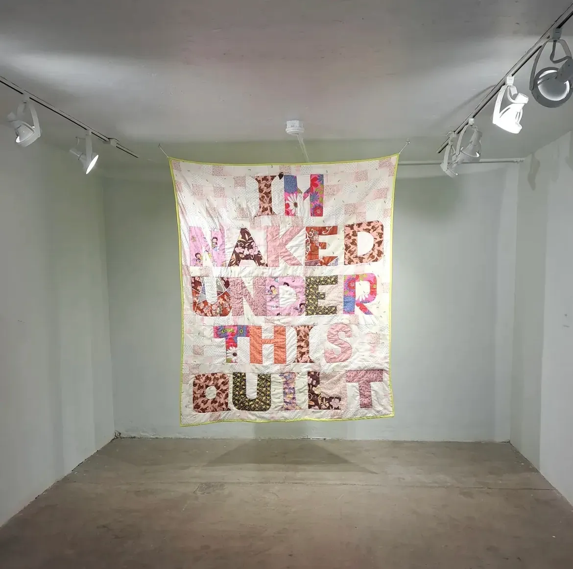 Large patchwork quilt hung from the ceiling of a white gallery space, reading "I'M NAKED UNDER THIS QUILT" in large letter forms cut from patterned fabrics — florals, animal prints, and mixed prints in pink, brown, and multicolor tones on a pale ground with a yellow-green border, illuminated by track lighting