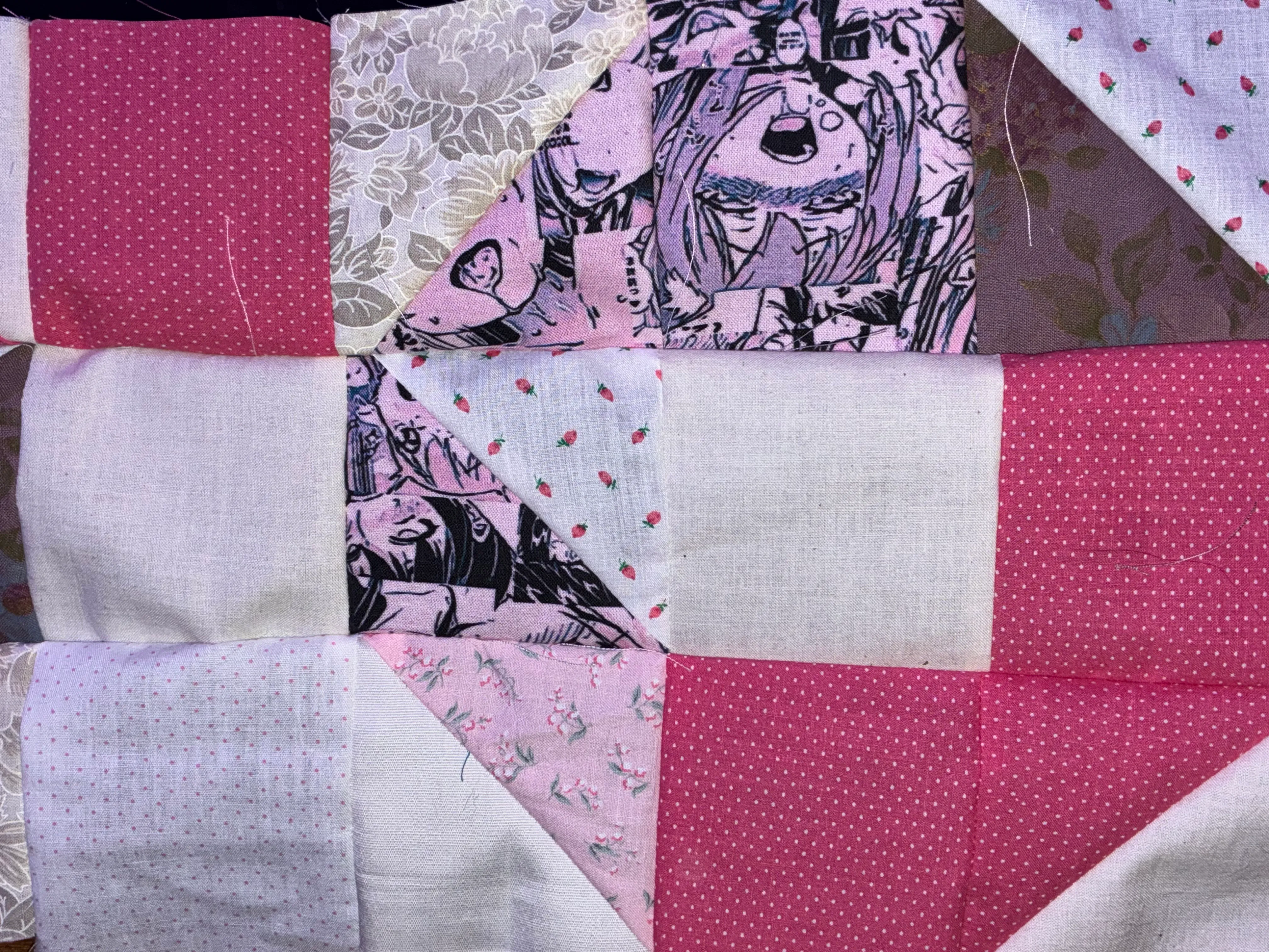 Close-up detail of a patchwork quilt top in progress, showing nine-patch style blocks in pink, white, and cream cotton fabrics including polka dots, custom anime-printed ahegao cotton, and floral prints, with raw edges visible at the seams
