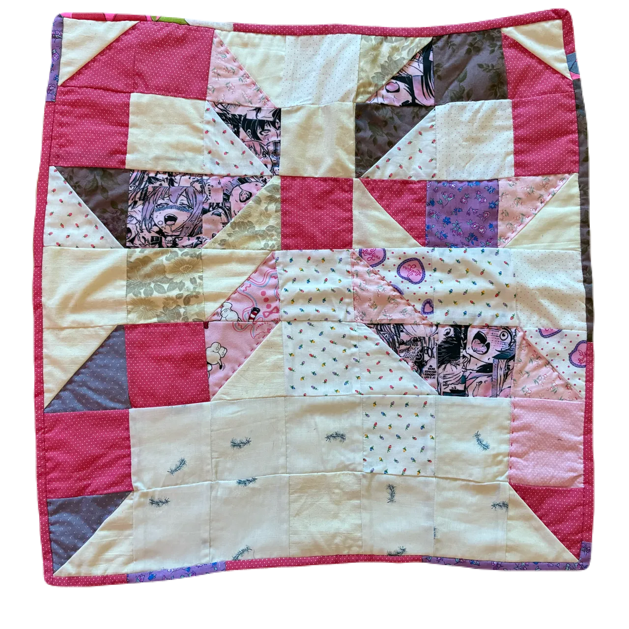 Small square patchwork quilt photographed against a black background, showing a large bow or pinwheel block formed by pink and cream triangles with anime-printed and polka-dot cotton fabrics, with a coral-pink border