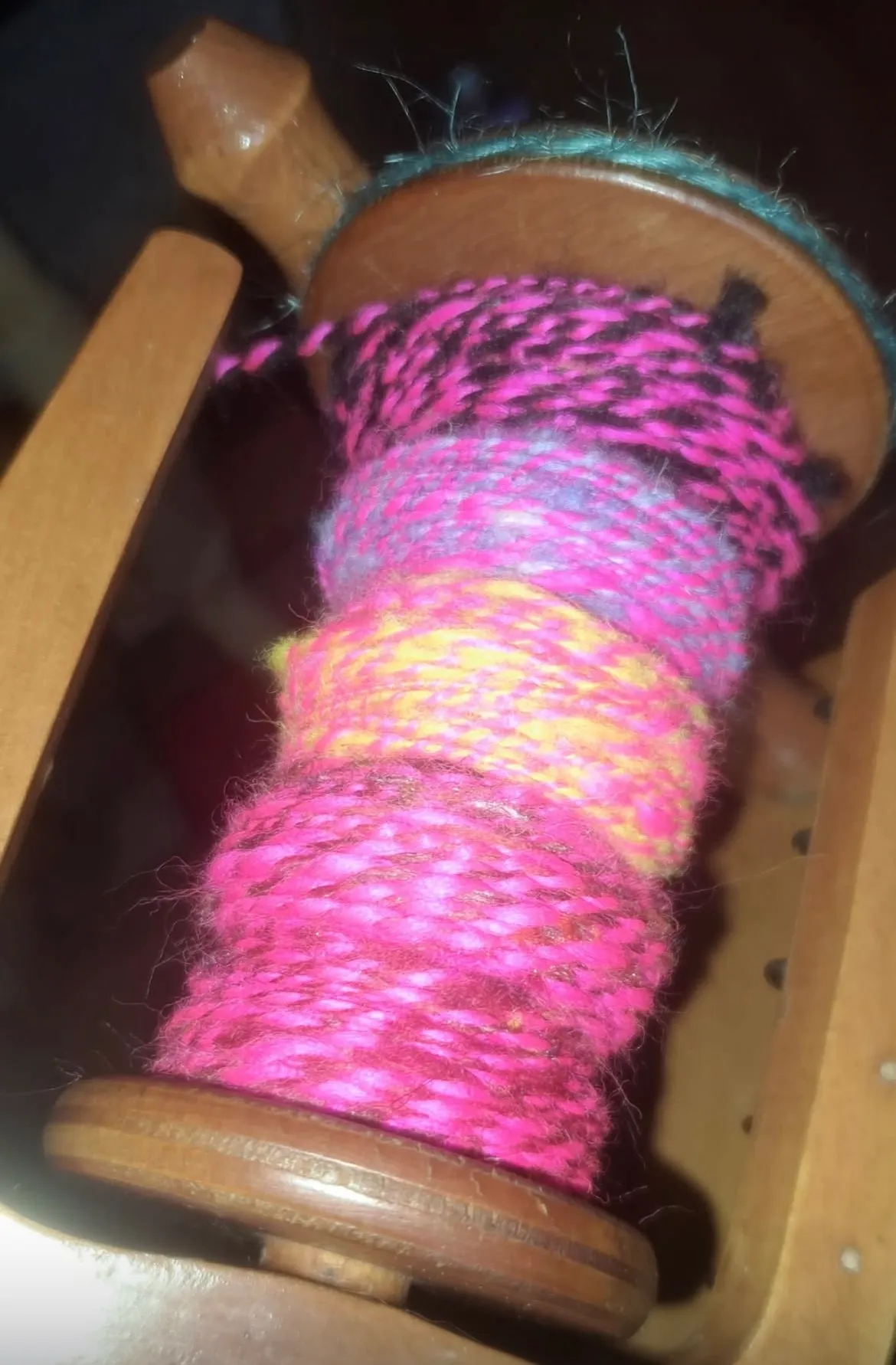 A wooden bobbin on a spinning wheel bobbin holder, wound with handspun wool yarn in distinct color bands — hot pink and black, grey-pink, yellow-orange, and deep rose — with a fluffy, textured surface