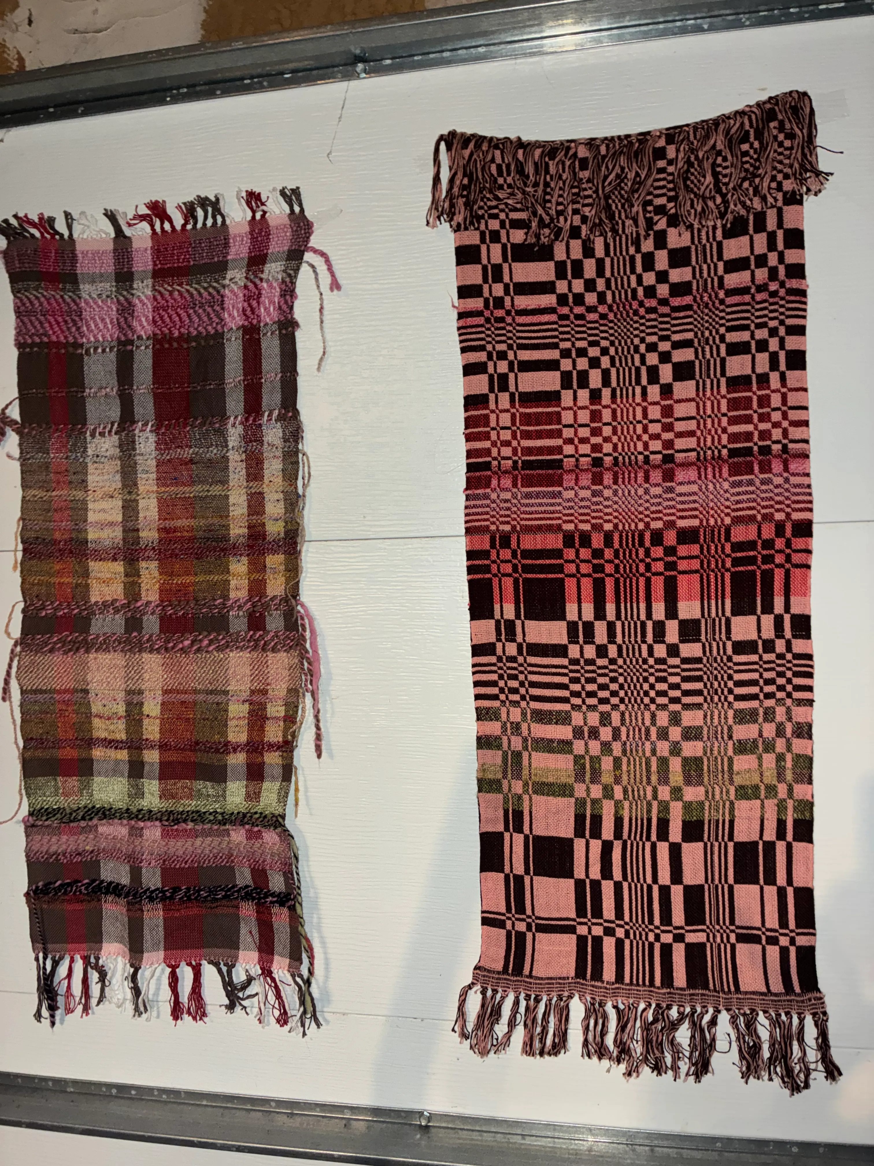 Two woven textiles displayed side by side on a white board, both with twisted fringe at the ends; the top piece shows a complex geometric pattern in pink and dark brown with alternating twill and plain weave sections; the bottom piece shows a plaid-like pattern in warm tones of pink, brown, olive, and burgundy