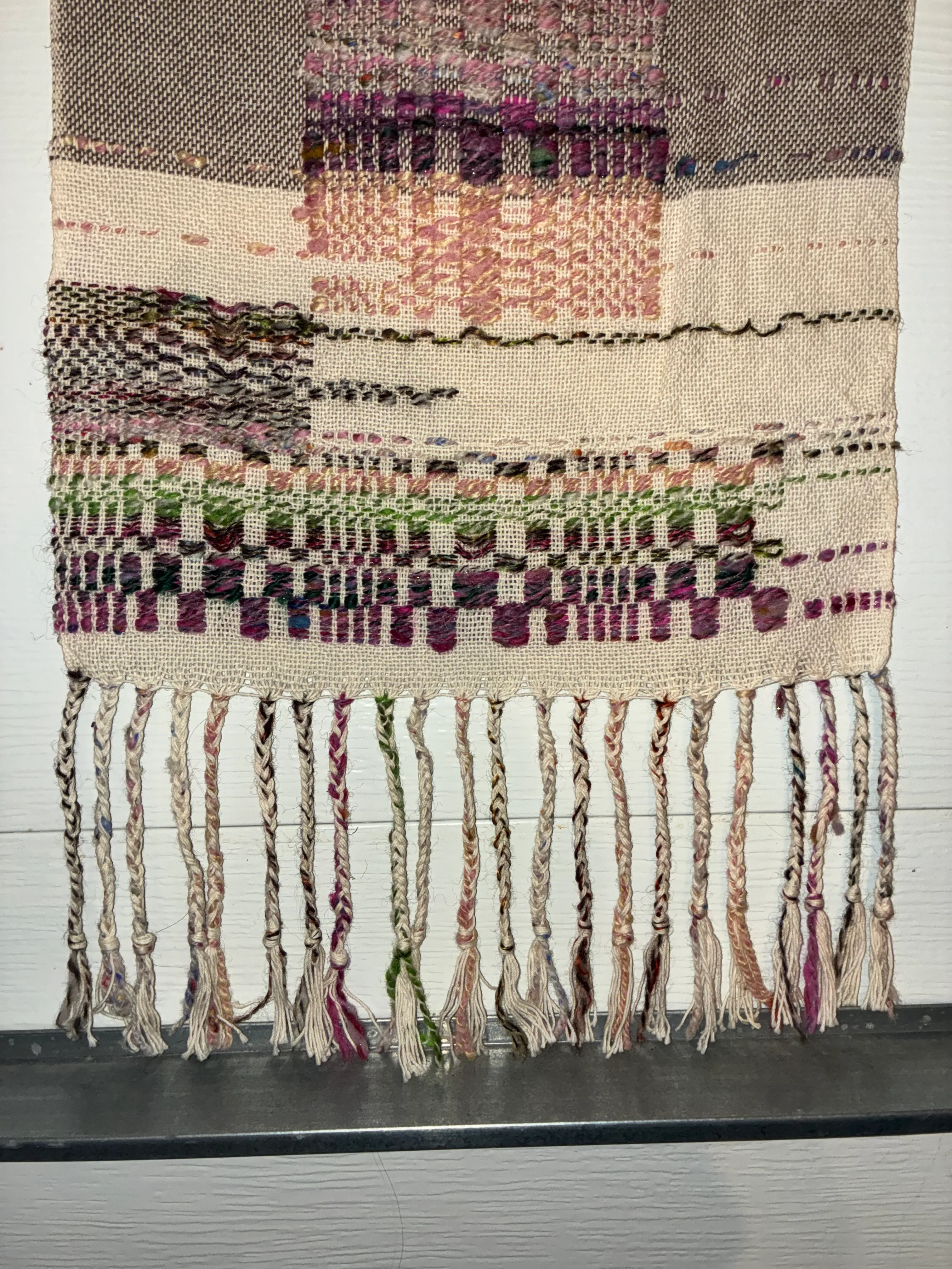 Close-up of the edge of a woven textile showing columns of overshot floats in purple, green, and pink handspun yarn against a cream cotton ground, with braided and twisted fringe in mixed natural and colored fibers