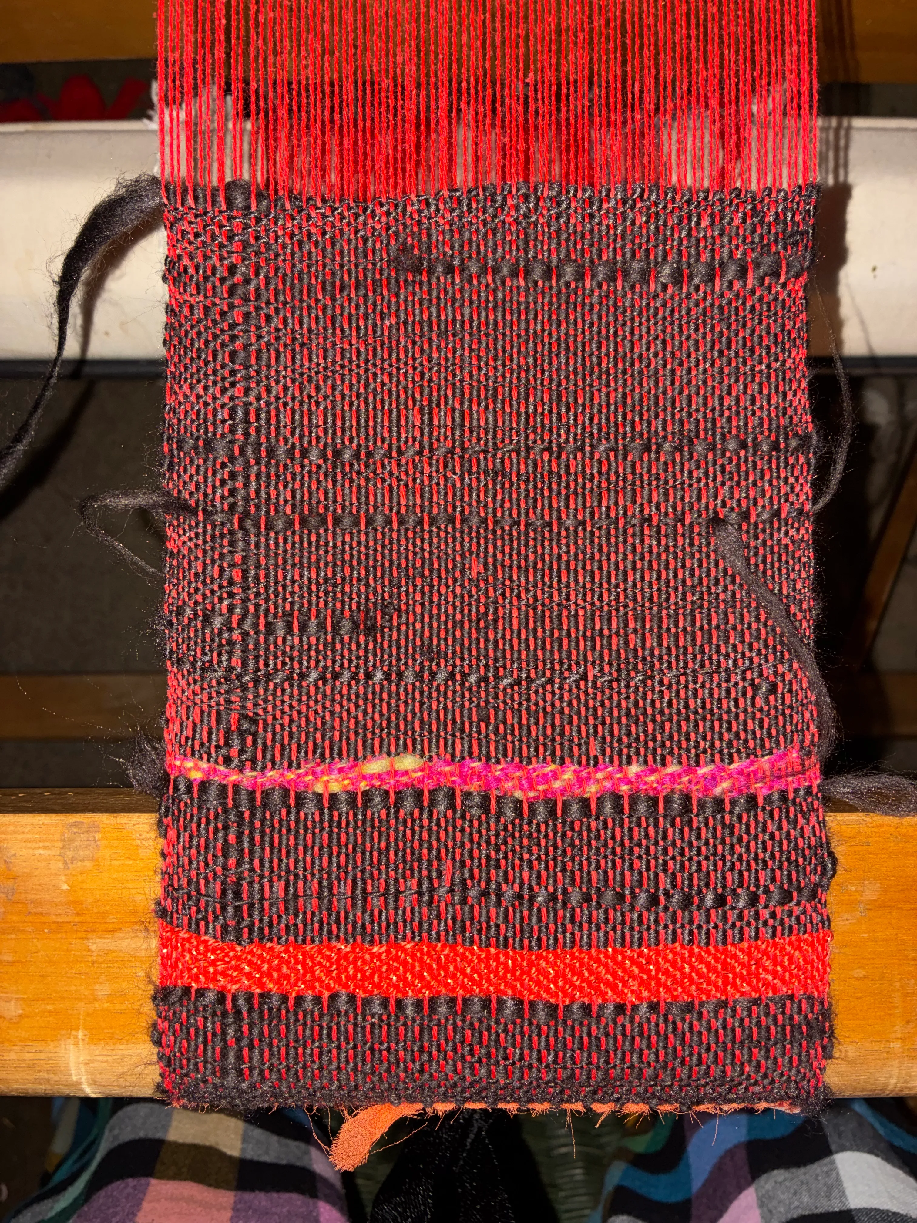 Close-up of woven textile on a loom, showing a bold pattern of red and black yarn with a horizontal pink stripe accent, still attached to the wooden loom beam