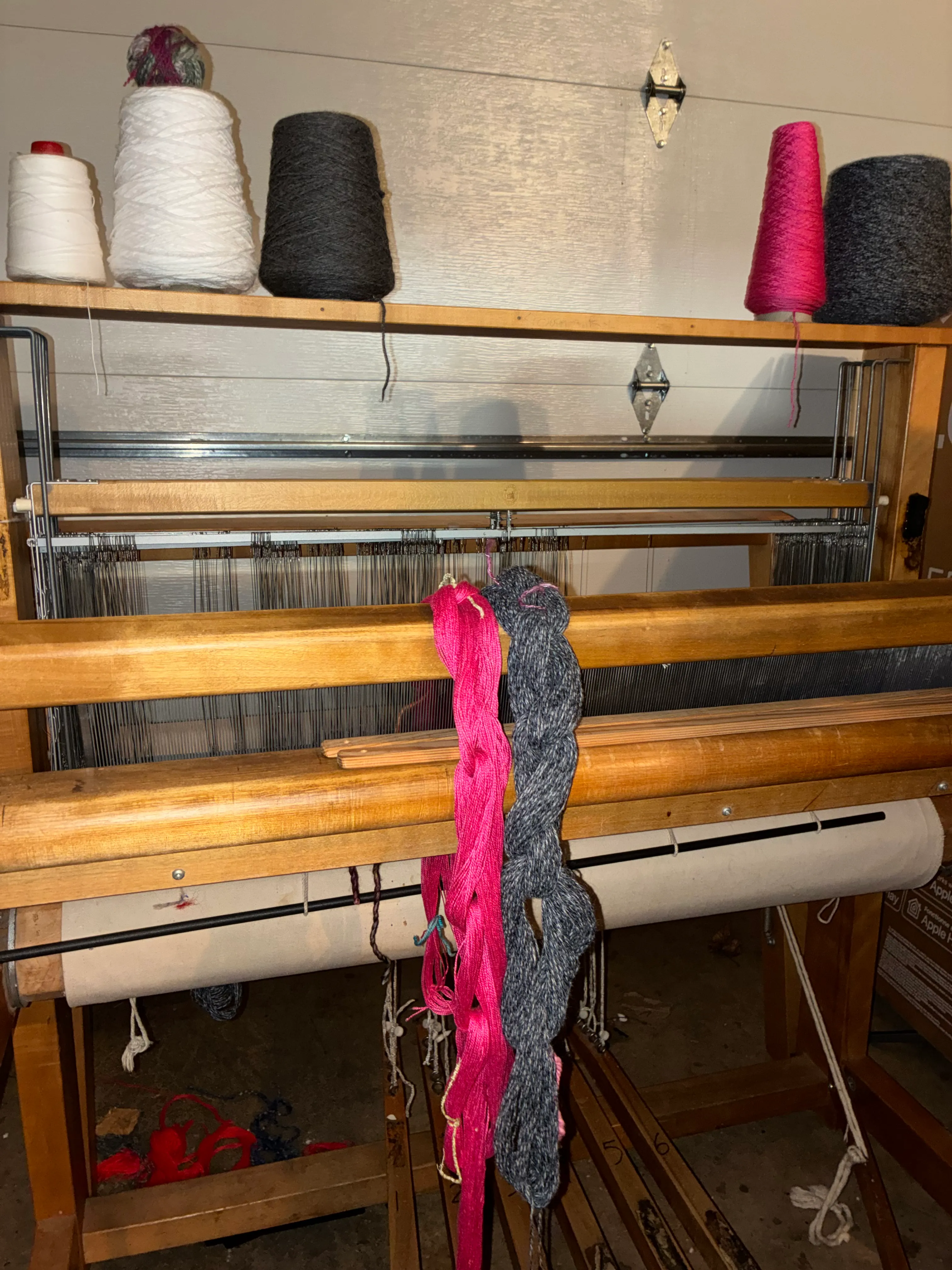 Wooden floor loom seen from above, with cones of pink, grey, and white yarn resting on the frame, heddles and warp threads visible, and a warp beam in the background