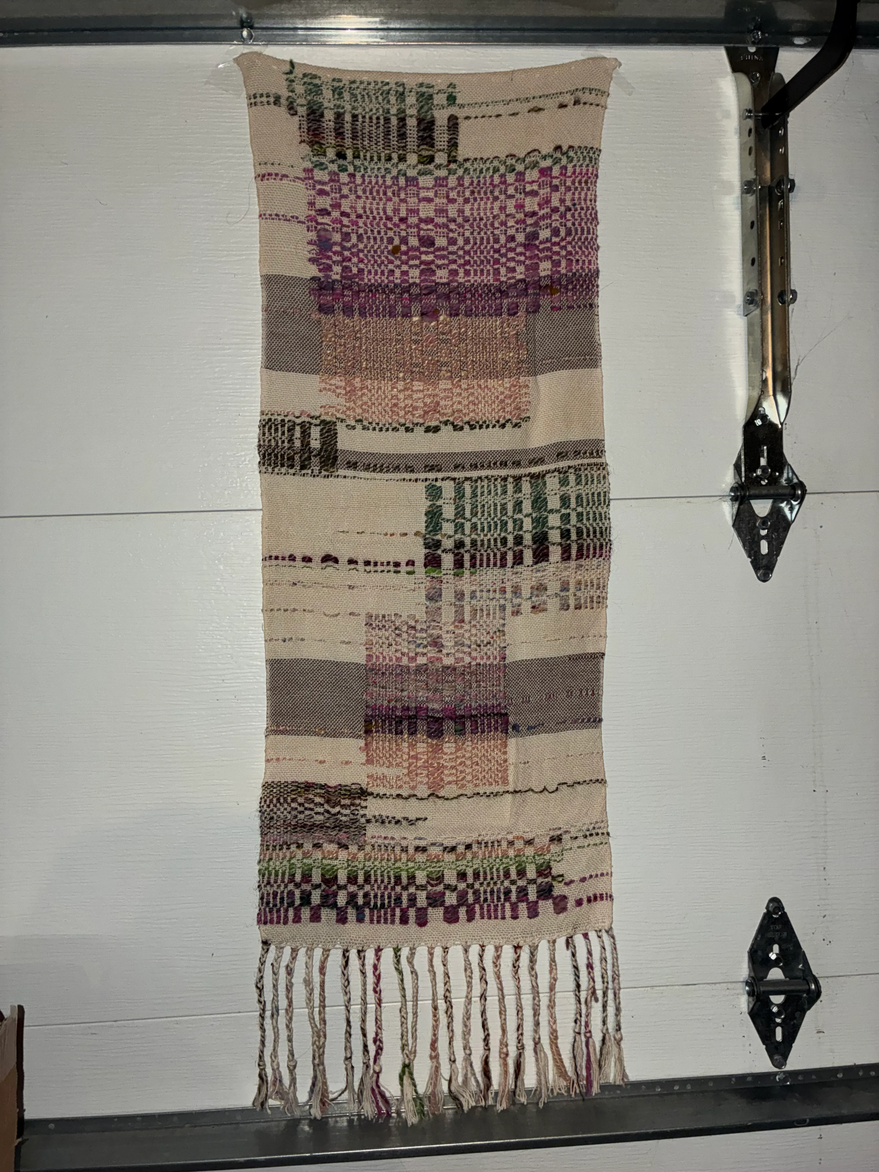 A long woven textile hung horizontally against a white wall, showing an overshot pattern with sections of cream plain weave interspersed with richly textured floats in purple, pink, green, and grey handspun yarn, with twisted fringe along the right edge