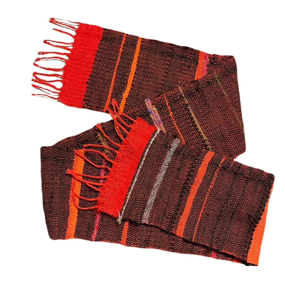 A woven scarf folded and laid diagonally against a white background, with a dark brown and deep red base and bold horizontal stripes of bright orange, with red knotted fringe on both short ends and a weaving shuttle tucked into the fold